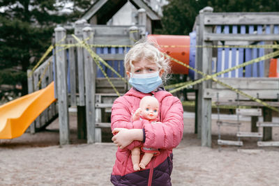 Sad caucasian girl in face mask with baby toy on closed playground outdoor. 