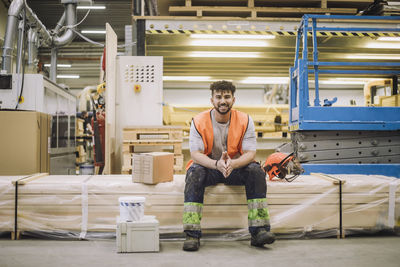Portrait of smiling young blue-collar worker sitting by hardhat in warehouse