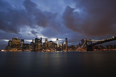 Illuminated brooklyn bridge over east river against cloudy sky at dusk