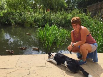 Woman crouching with dog on retaining wall by lake on sunny day