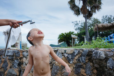 Cropped hand of young man showering shirtless boy while standing outdoors