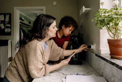 Mother charging smart phone at kitchen counter by son at home