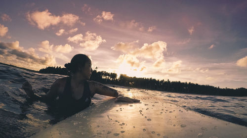 Woman swimming with surfboard in lake against sky during sunset