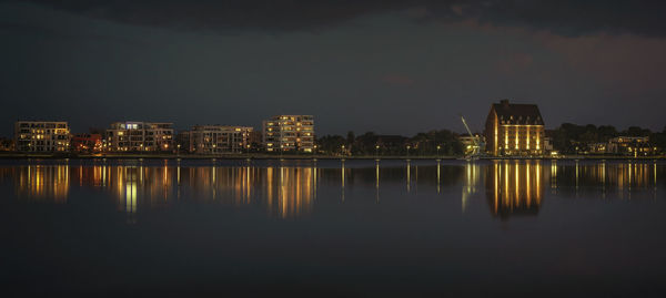 Illuminated buildings by lake against sky in city at night