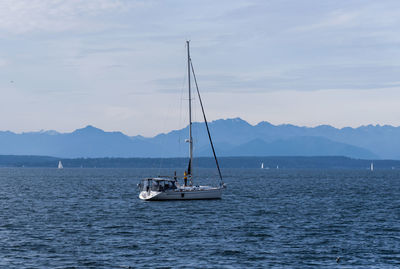 Sailboat off shore mid day sun with olympic mountains and people standing on deck.