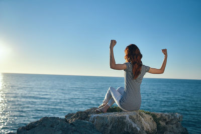 Rear view of woman flexing muscles while sitting on rock against sea