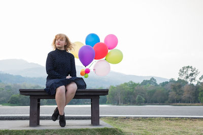 Full length of woman with balloons sitting on bench against sky