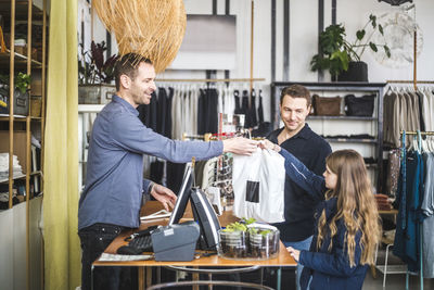 Smiling cashier giving shopping bag to girl by father in boutique