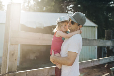 A father holds his young daughter at a farm on a sunny evening.