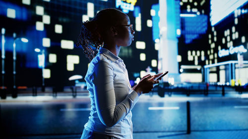 Side view of woman using mobile phone while standing in city at night