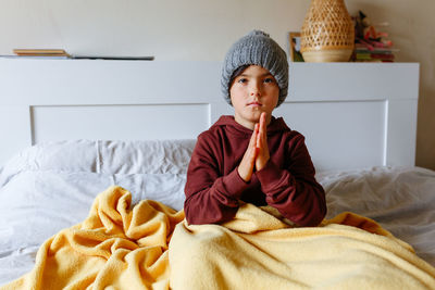 Girl wearing grey warm hat sits on the bed and rub hands