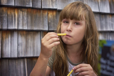 Portrait of girl eating french fries against wooden wall