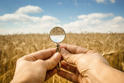 Cropped hands holding magnifying glass on agricultural field against sky