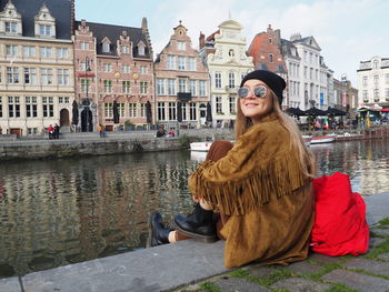 Portrait of smiling woman sitting by canal against buildings