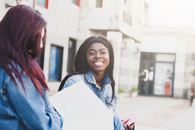 Smiling woman with books sitting by friend against building 