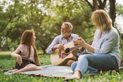 Happy grandfather playing guitar by granddaughter and woman in park