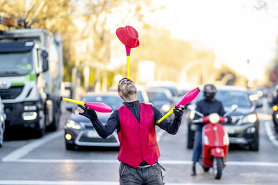 Performer balancing juggling pin and hat on nose in front of cars