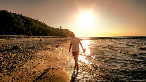 Man walking on shore at beach against sky during sunset