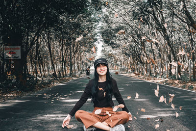 Portrait of smiling young woman throwing leaves while sitting on road amidst trees in forest