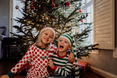 Siblings in costume sitting against christmas tree at home