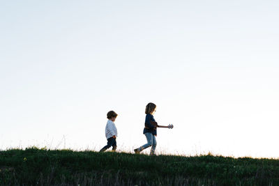 From below side view of little girl with guitar followed by younger brother walking in green field in summer evening in countryside