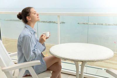 Beautiful young woman holding tea cup while sitting in balcony against sea