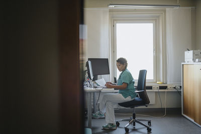 Side view of female doctor checking medical record while sitting at desk in office