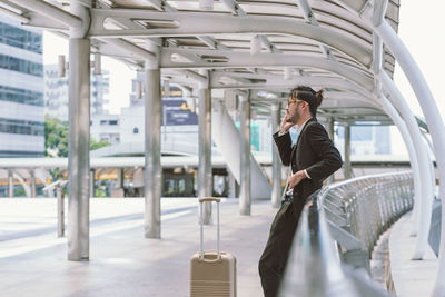 Businessman with luggage talking through phone on footbridge in city