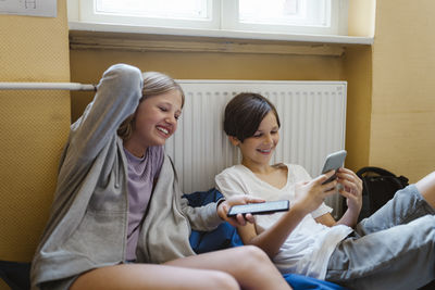 Happy male and female students using smart phone while sitting near heater at school