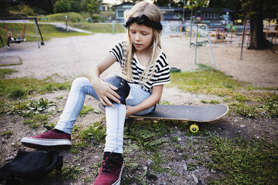 Girl tying kneepad while sitting on skateboard at park