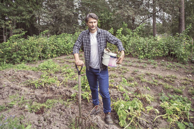 Full length portrait of confident man with gardening fork and bucket at organic farm