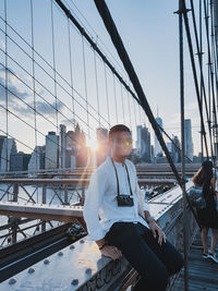 Man sitting on bridge against sky in city