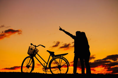 Silhouette person with bicycle by woman standing on field against sky during sunset