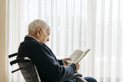 Senior man sitting in wheelchair reading a book near the window