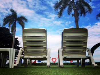 Empty chairs and palm trees in yard against sky