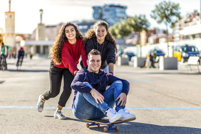 Portrait of happy teenage girls pushing friend sitting on skateboard 