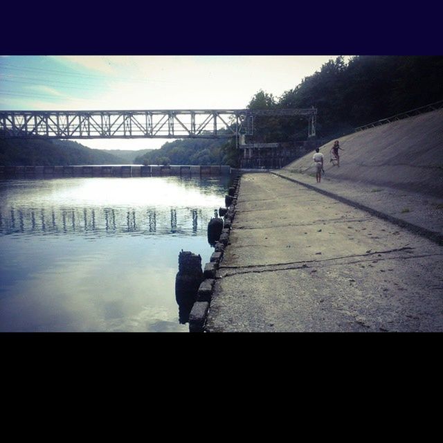 water, silhouette, built structure, architecture, bridge - man made structure, connection, men, transfer print, river, lifestyles, auto post production filter, walking, sky, bridge, railing, reflection, transportation, full length