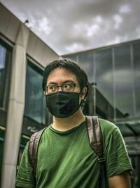 Portrait of young man standing outdoors against buildings and cloudy sky.