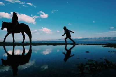 Boy following man riding horse by song kol lake against blue sky