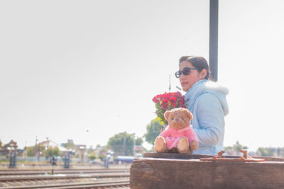 Woman with bouquet and teddy bear sitting at railroad station platform during sunny day