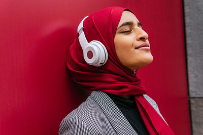 Side view of dreamy arab female in wireless headphones and traditional hijab standing near urban red building and enjoying music with closed eyes