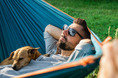 Smiling attractive european man in sunglasses is resting in hammock with his cute little dog.