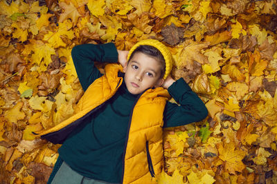 A boy child in a yellow jacket and hat lies on autumn leaves in the forest