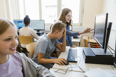 Girl pointing on monitor screen with male friend sitting at desk in classroom at school