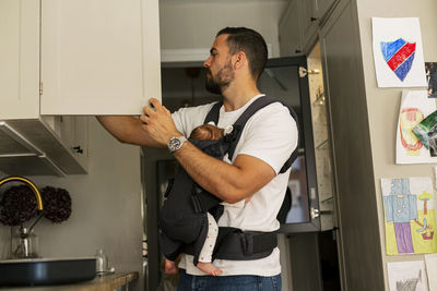 Father opening cabinet while carrying son in kitchen at home