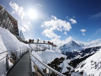 People walking on footbridge by snow covered mountains against sky