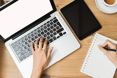 Cropped hands of businessman writing on book while using laptop at desk