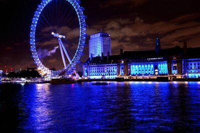 Illuminated ferris wheel by river against sky at night