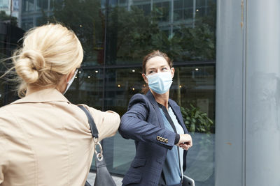 Businesswoman and colleague with face mask greeting with elbow bump while standing against office building