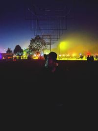 Silhouette people sitting by illuminated lights against sky at night